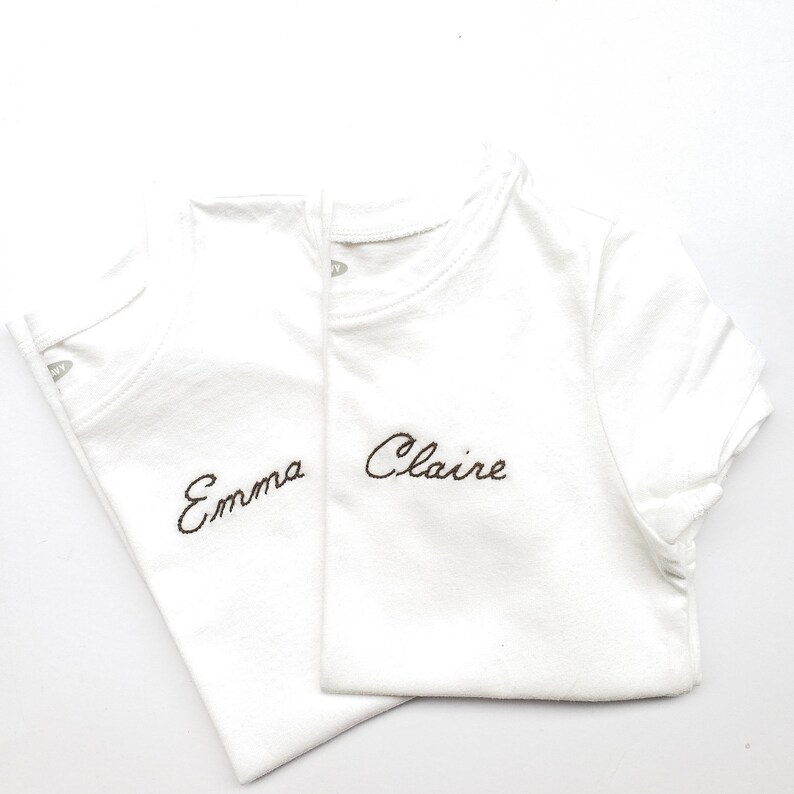 May include: Two white t-shirts with brown embroidered names. The top shirt says "Claire" and the bottom shirt says "Emma".