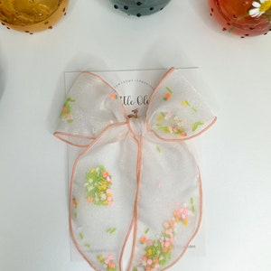 Op de afbeelding: Een witte, doorschijnende strik met een perzikkleurige rand en een bloemenpatroon. De strik is in het midden geknoopt en heeft een klein label met de tekst "Little Ole".