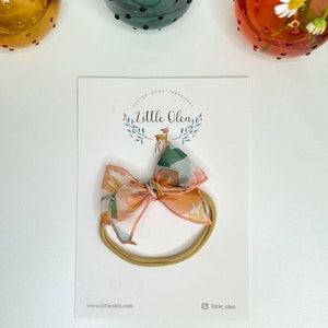 Op de afbeelding: Een nude elastisch hoofdband met een strik in een perzik- en groene bloemenprint. Het hoofdband wordt getoond op een witte kaart met de tekst "Little Olen" en het websiteadres "www.littleolen.com".
