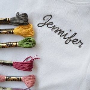 May include: A white t-shirt with the name "Jennifer" embroidered in black thread.  Six spools of embroidery floss in various colors are arranged on the left side of the image.