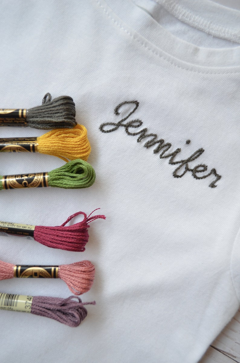 May include: A white t-shirt with the name "Jennifer" embroidered in black thread.  Six spools of embroidery floss in various colors are arranged on the left side of the image.