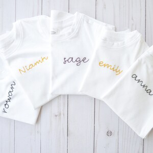May include: Five white t-shirts with different names embroidered on them in various colors. The names are "Rowan", "Niamh", "Sage", "Emily", and "Anna".