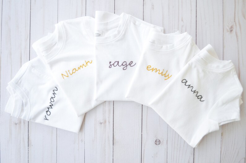 May include: Five white t-shirts with different names embroidered on them in various colors. The names are "Rowan", "Niamh", "Sage", "Emily", and "Anna".