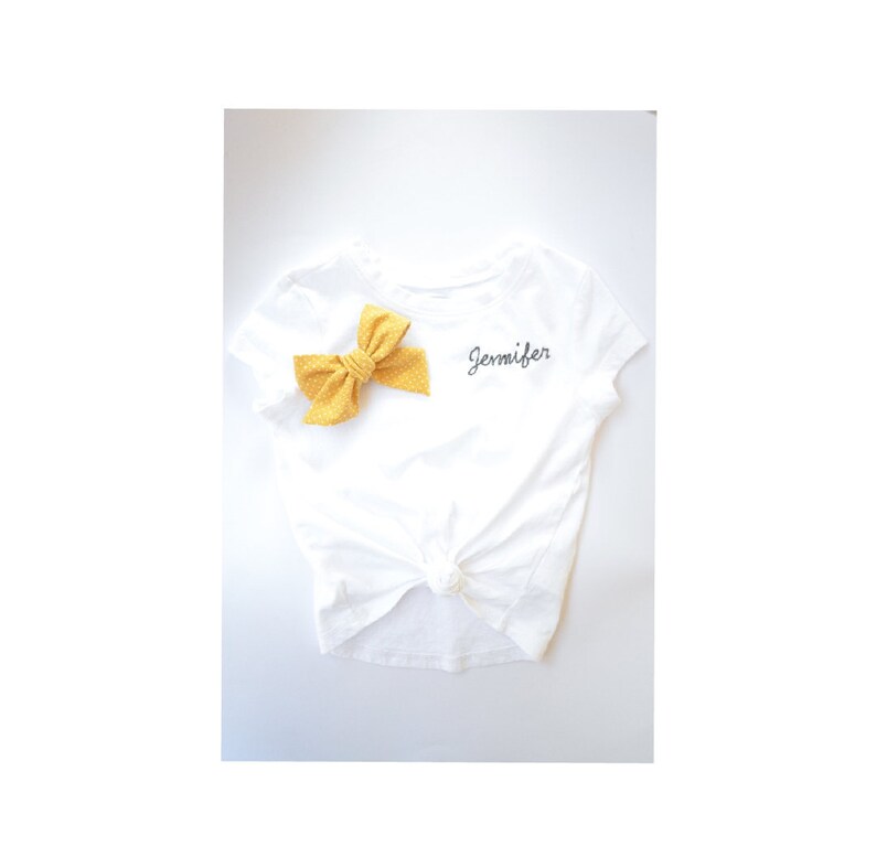 May include: White t-shirt with a yellow bow and the name "Jennifer" embroidered on the front.