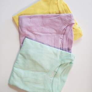 May include: Three plain t-shirts in pastel colors: yellow, lavender, and light green. The shirts are folded and stacked on top of each other.
