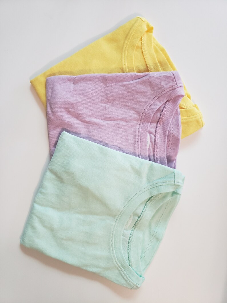 May include: Three plain t-shirts in pastel colors: yellow, lavender, and light green. The shirts are folded and stacked on top of each other.