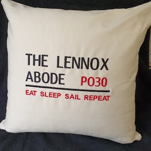 May include: Cream-coloured square cushion with black text reading "THE LENNOX ABODE P030" and red text below saying "EAT SLEEP SAIL REPEAT". The cushion is on a dark blue surface.