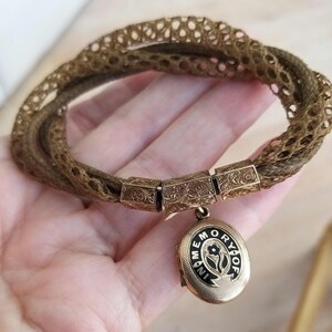 May include: A vintage bracelet featuring woven brown strands and ornate gold-toned rectangular links. A small, oval locket with the words "IN MEMORY OF" and a floral design hangs from the bracelet, showcasing a classic, antique aesthetic.