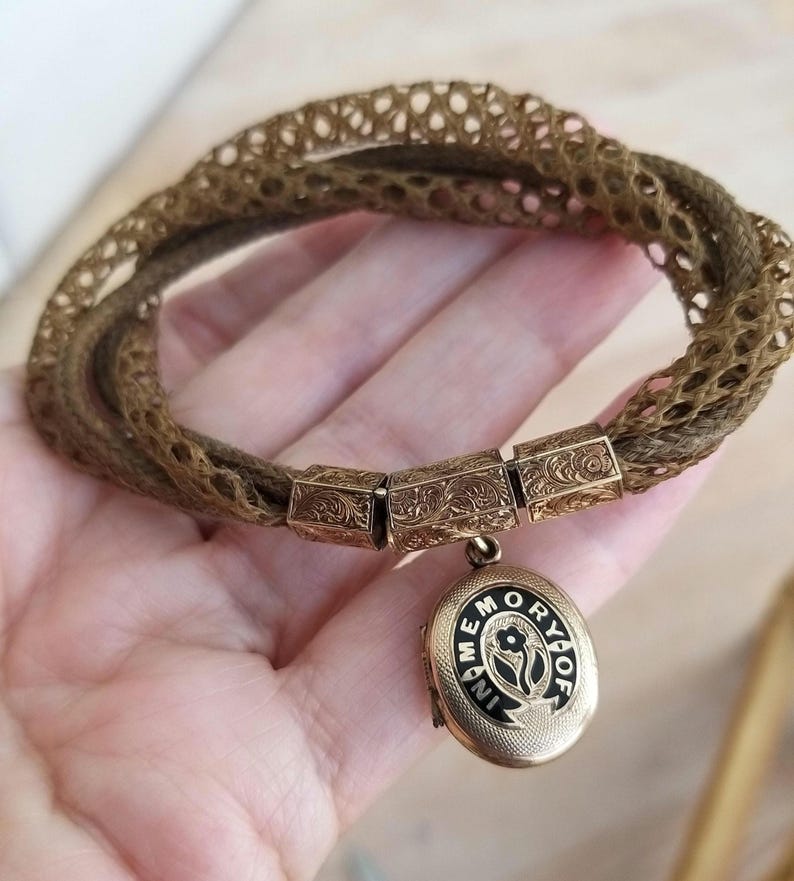 May include: A vintage bracelet featuring woven brown strands and ornate gold-toned rectangular links. A small, oval locket with the words "IN MEMORY OF" and a floral design hangs from the bracelet, showcasing a classic, antique aesthetic.