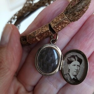 May include: Antique gold-tone locket pendant with a woven hair compartment and a small portrait inside. The locket is attached to a braided hair bracelet. The locket is oval-shaped and has an ornate design.
