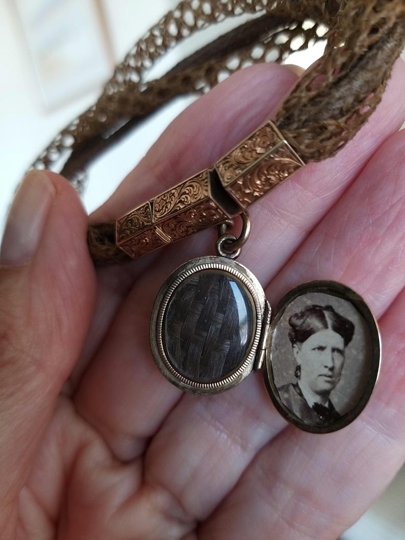 May include: Antique gold-tone locket pendant with a woven hair compartment and a small portrait inside. The locket is attached to a braided hair bracelet. The locket is oval-shaped and has an ornate design.