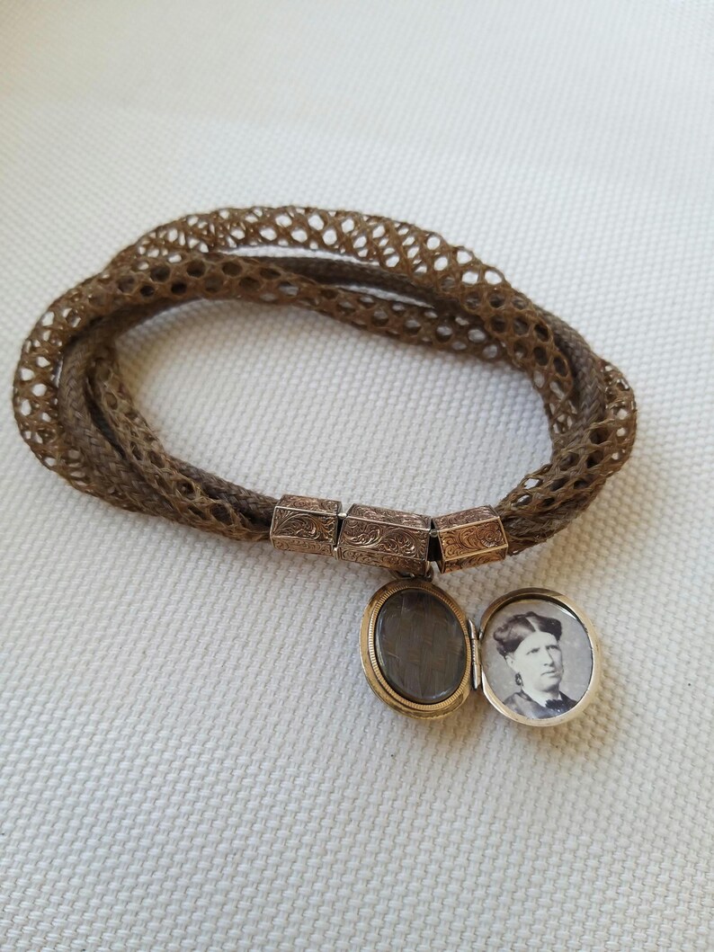 May include: A vintage brown hair bracelet with a woven design. The bracelet features ornate gold-toned metal clasps and a small locket with a portrait inside. The locket is open, revealing the image.