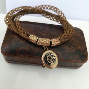 May include: A vintage brown woven bracelet with gold-toned rectangular accents and a small oval locket. The locket has black enamel detailing and the words "In Memory Of". The bracelet rests on a brown leather box.
