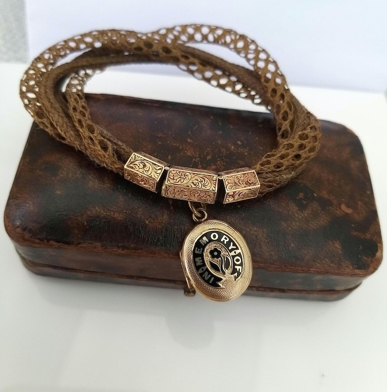 May include: A vintage brown woven bracelet with gold-toned rectangular accents and a small oval locket. The locket has black enamel detailing and the words "In Memory Of". The bracelet rests on a brown leather box.