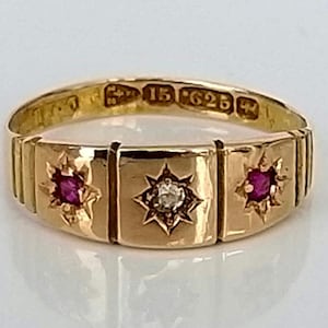 Könnte beinhalten: Ein Goldring mit drei rechteckigen Abschnitten, die jeweils ein Sternmuster aufweisen. Der mittlere Abschnitt hat einen weißen Stein, während die beiden anderen Abschnitte rote Steine haben.