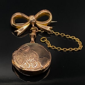Op de afbeelding: Een gouden medaillon met een delicate strik en een ketting. Het medaillon heeft een ingewikkeld bloemmotief aan de voorkant.