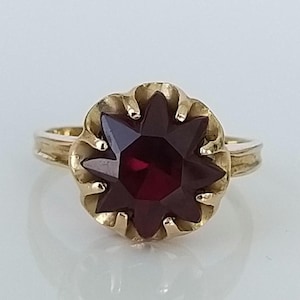 May include: A gold ring featuring a large, faceted, dark red gemstone set in a flower-shaped setting.