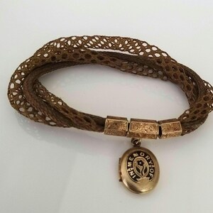 May include: A vintage brown hair bracelet with a woven design. The bracelet features gold-toned rectangular clasps and a small, round locket pendant with the inscription "IN MEMORY OF". The locket has a floral design.