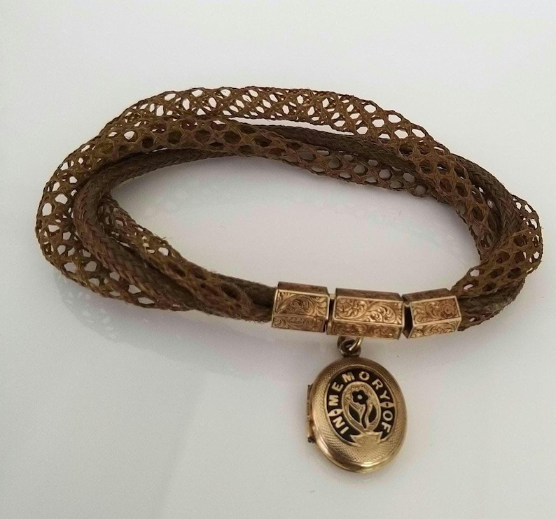 May include: A vintage brown hair bracelet with a woven design. The bracelet features gold-toned rectangular clasps and a small, round locket pendant with the inscription "IN MEMORY OF". The locket has a floral design.