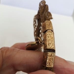 May include: A gold-toned antique bracelet with ornate, engraved rectangular links. The bracelet is attached to a woven, brown cord. A small, round, gold-toned locket hangs from the bracelet.