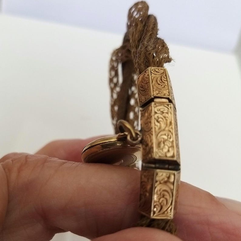 May include: A gold-toned antique bracelet with ornate, engraved rectangular links. The bracelet is attached to a woven, brown cord. A small, round, gold-toned locket hangs from the bracelet.