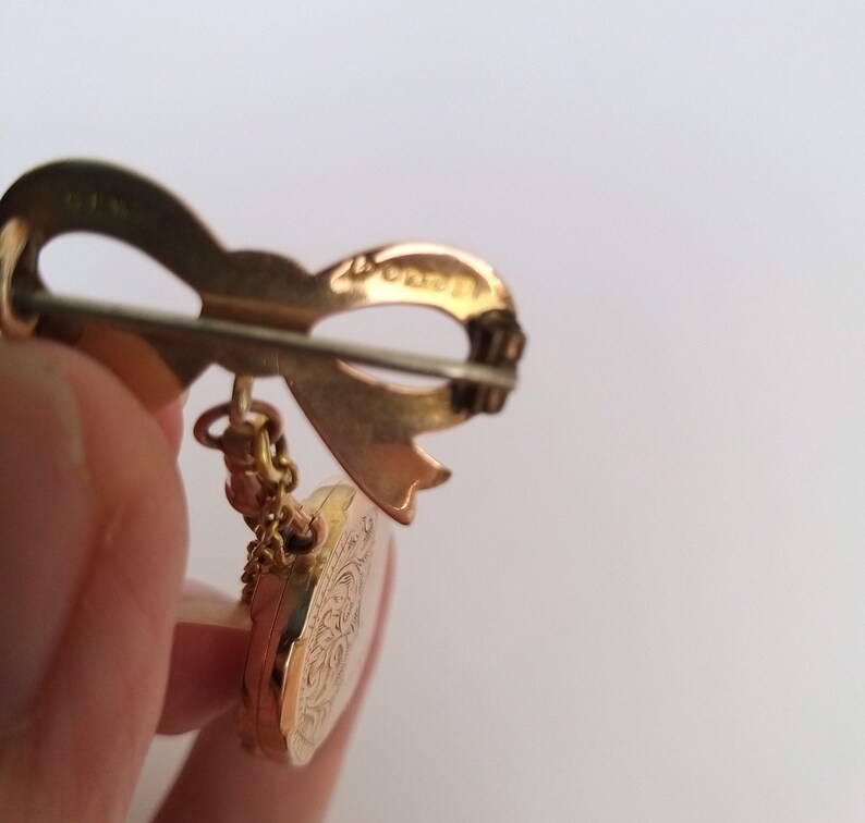 Op de afbeelding: Gouden broche met een strik en een rond medaillon met een bloemmotief. De achterkant van de broche heeft de inscriptie "14K Gold Filled".