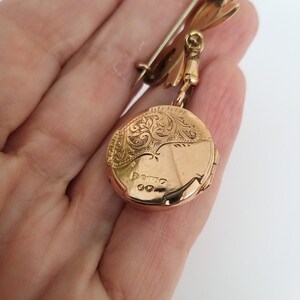 Op de afbeelding: Een gouden medaillon met een gegraveerd bloemenmotief en een scharnierende opening. Het medaillon is bevestigd aan een gouden ketting met een veiligheidsspeld.