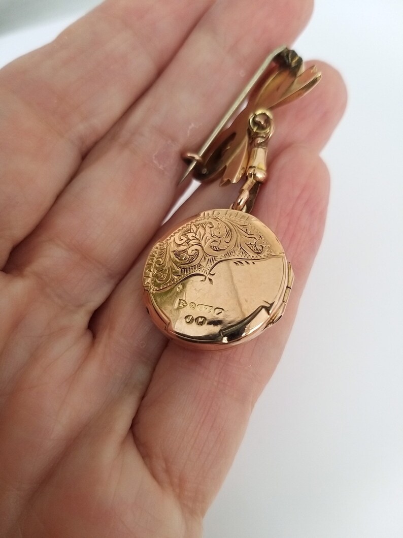 Op de afbeelding: Een gouden medaillon met een gegraveerd bloemenmotief en een scharnierende opening. Het medaillon is bevestigd aan een gouden ketting met een veiligheidsspeld.