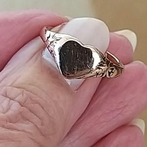 May include: A silver heart-shaped ring with floral detailing on the band. The ring is worn on a finger.