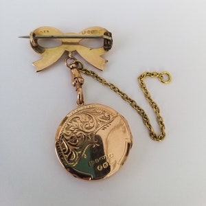 Op de afbeelding: Een gouden hanger met een gedetailleerd bloemenmotief en een strik-vormige broche bevestigd aan een ketting. De hanger heeft de inscriptie "DOCTA" op de achterkant.