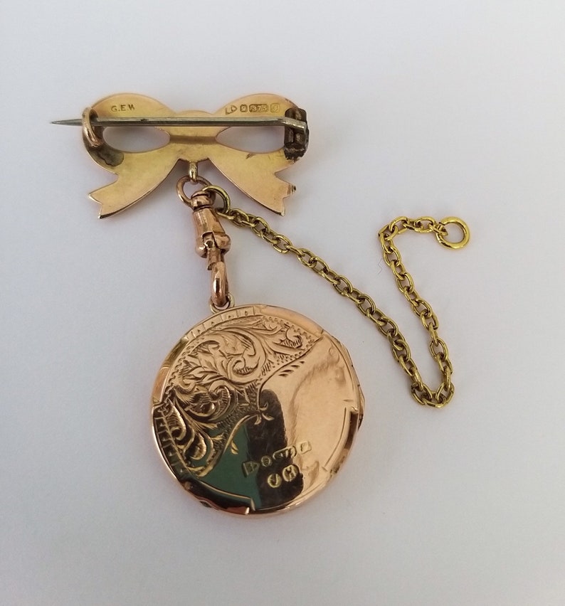 Op de afbeelding: Een gouden hanger met een gedetailleerd bloemenmotief en een strik-vormige broche bevestigd aan een ketting. De hanger heeft de inscriptie "DOCTA" op de achterkant.