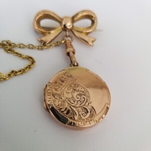 Op de afbeelding: Een gouden medaillon met een delicate ketting en een speld in de vorm van een strik. Het medaillon is gegraveerd met een bloemmotief.