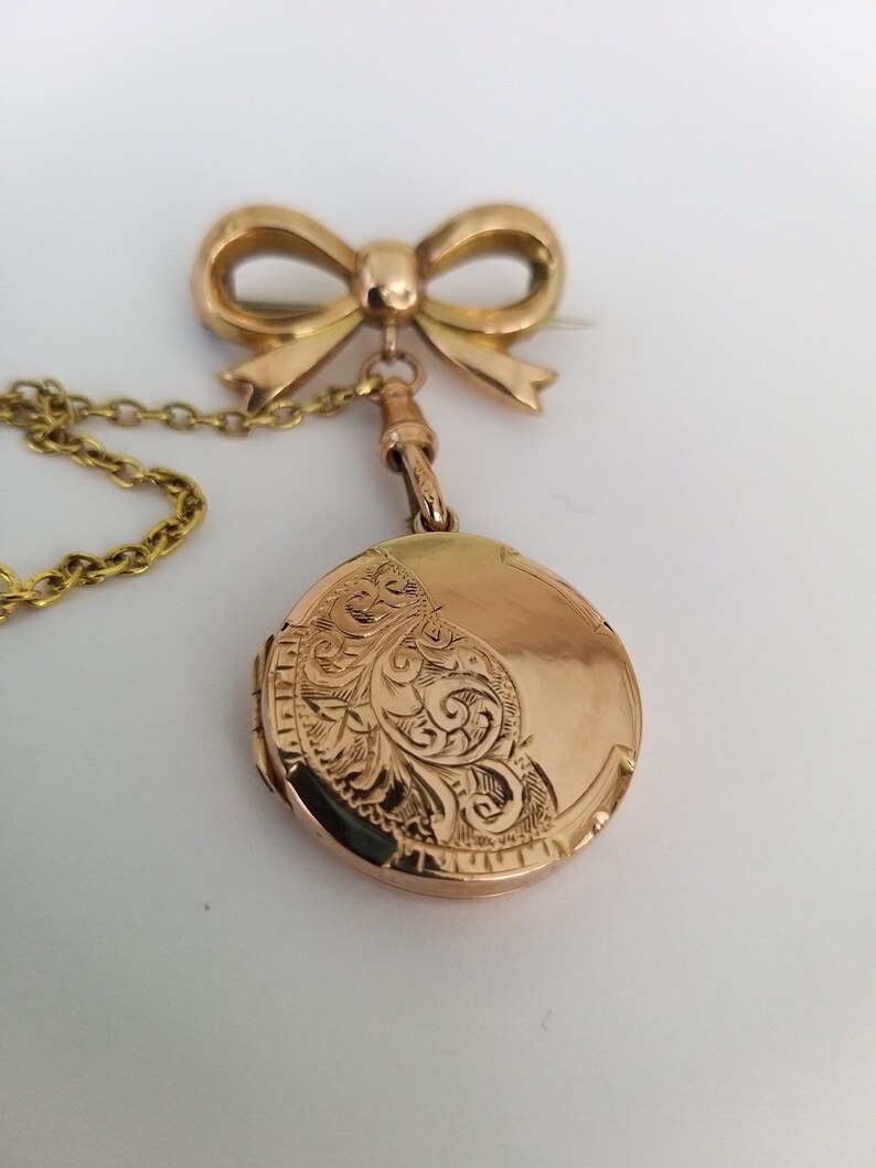 Op de afbeelding: Een gouden medaillon met een delicate ketting en een speld in de vorm van een strik. Het medaillon is gegraveerd met een bloemmotief.