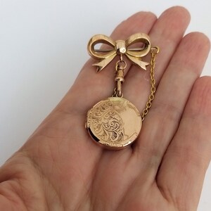 Op de afbeelding: Een gouden medaillon met een delicate strikbedel en een ketting. Het medaillon heeft een ingegraveerd ontwerp.