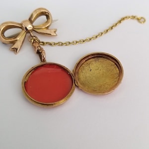 Op de afbeelding: Een gouden medaillon met een rode binnenkant en een ketting in de vorm van een strik eraan bevestigd. Het medaillon is open, waardoor de rode binnenkant zichtbaar is.