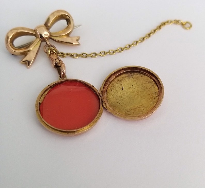 Op de afbeelding: Een gouden medaillon met een rode binnenkant en een ketting in de vorm van een strik eraan bevestigd. Het medaillon is open, waardoor de rode binnenkant zichtbaar is.