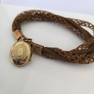 May include: A vintage brown hair bracelet with a gold locket. The locket is oval-shaped with an engraved design and opens to reveal space for a photo. The bracelet is made of woven brown hair and has gold connectors.