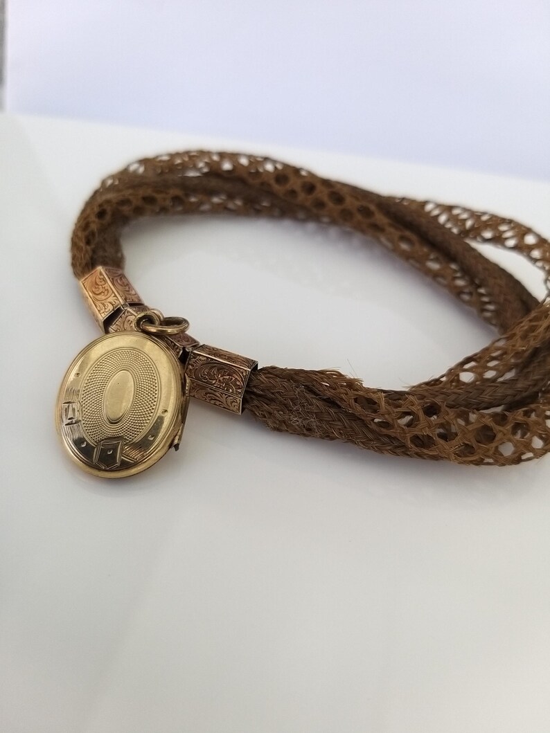 May include: A vintage brown hair bracelet with a gold locket. The locket is oval-shaped with an engraved design and opens to reveal space for a photo. The bracelet is made of woven brown hair and has gold connectors.