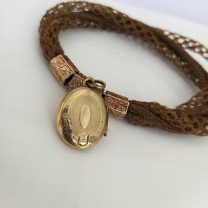 May include: A vintage gold locket with an oval design, attached to a woven brown hair bracelet. The locket is suspended from decorative gold connectors. The bracelet is made of tightly woven brown hair.