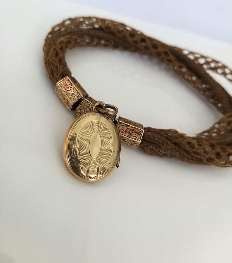 May include: A vintage gold locket with an oval design, attached to a woven brown hair bracelet. The locket is suspended from decorative gold connectors. The bracelet is made of tightly woven brown hair.