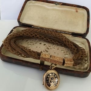 May include: An antique gold locket with the words "In Memory Of" and a floral design, attached to a woven hair bracelet. The bracelet is displayed in a vintage brown leather box with a cream-colored interior.