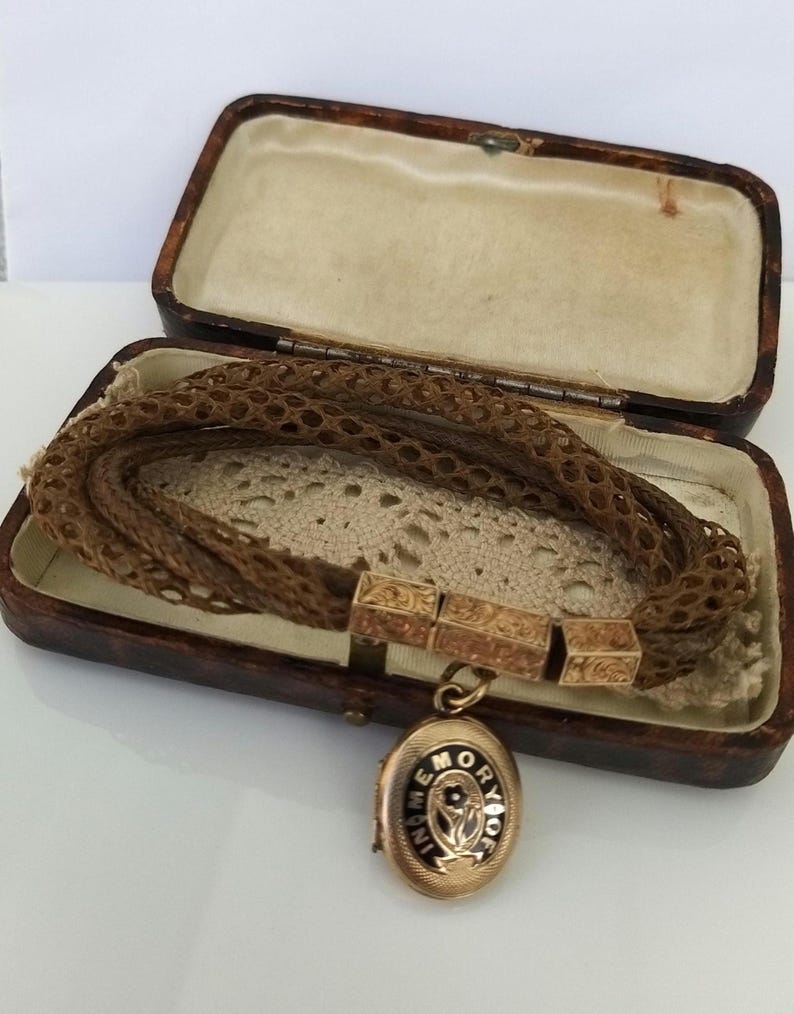 May include: An antique gold locket with the words "In Memory Of" and a floral design, attached to a woven hair bracelet. The bracelet is displayed in a vintage brown leather box with a cream-colored interior.