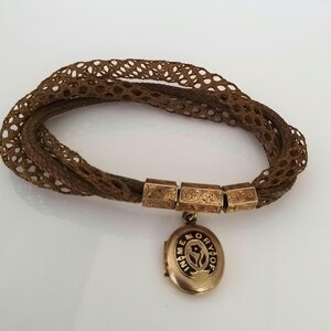 May include: A brown woven hair bracelet with gold-toned rectangular connectors and a round locket pendant. The locket is inscribed with the words "IN MEMORY OF" and a floral design. The bracelet is a vintage piece.