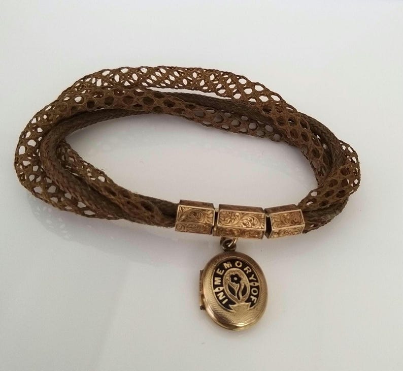 May include: A brown woven hair bracelet with gold-toned rectangular connectors and a round locket pendant. The locket is inscribed with the words "IN MEMORY OF" and a floral design. The bracelet is a vintage piece.