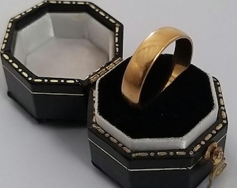 May include: A gold wedding band in a black ring box with a white interior. The box has a decorative pattern around the edge.