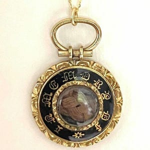 Op de afbeelding: Een gouden ketting met een hanger met een zwarte emaille rand en een glazen inzetstuk met een lok haar. De hanger is gegraveerd met de letters "H.S.M.E.R.D."