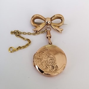 Op de afbeelding: Een gouden medaillon met een delicate strikbedel en een ketting. Het medaillon is gegraveerd met een bloemmotief.