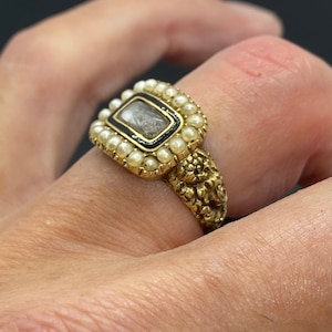 Op de afbeelding: Een gouden ring met een rechthoekige steen gezet in een zwart emaille frame. De steen is omringd door een rij parels. De band van de ring is versierd met een textuurontwerp.