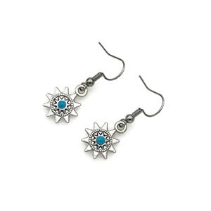 May include: A pair of silver-toned earrings featuring a sunburst design with a turquoise-colored center. The earrings have a hook closure and are displayed against a white background. The earrings are a jewelry item.