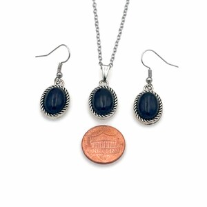 May include: A silver necklace and earrings set with black oval stones. The pendant and earrings are surrounded by a silver rope design. The set is shown next to a US penny for size comparison.